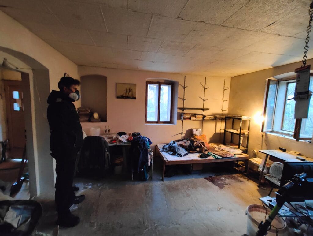 Muž v ochranné masce při vyklízení nepořádného sklepa s rozestavěným nábytkem a osvětlením.