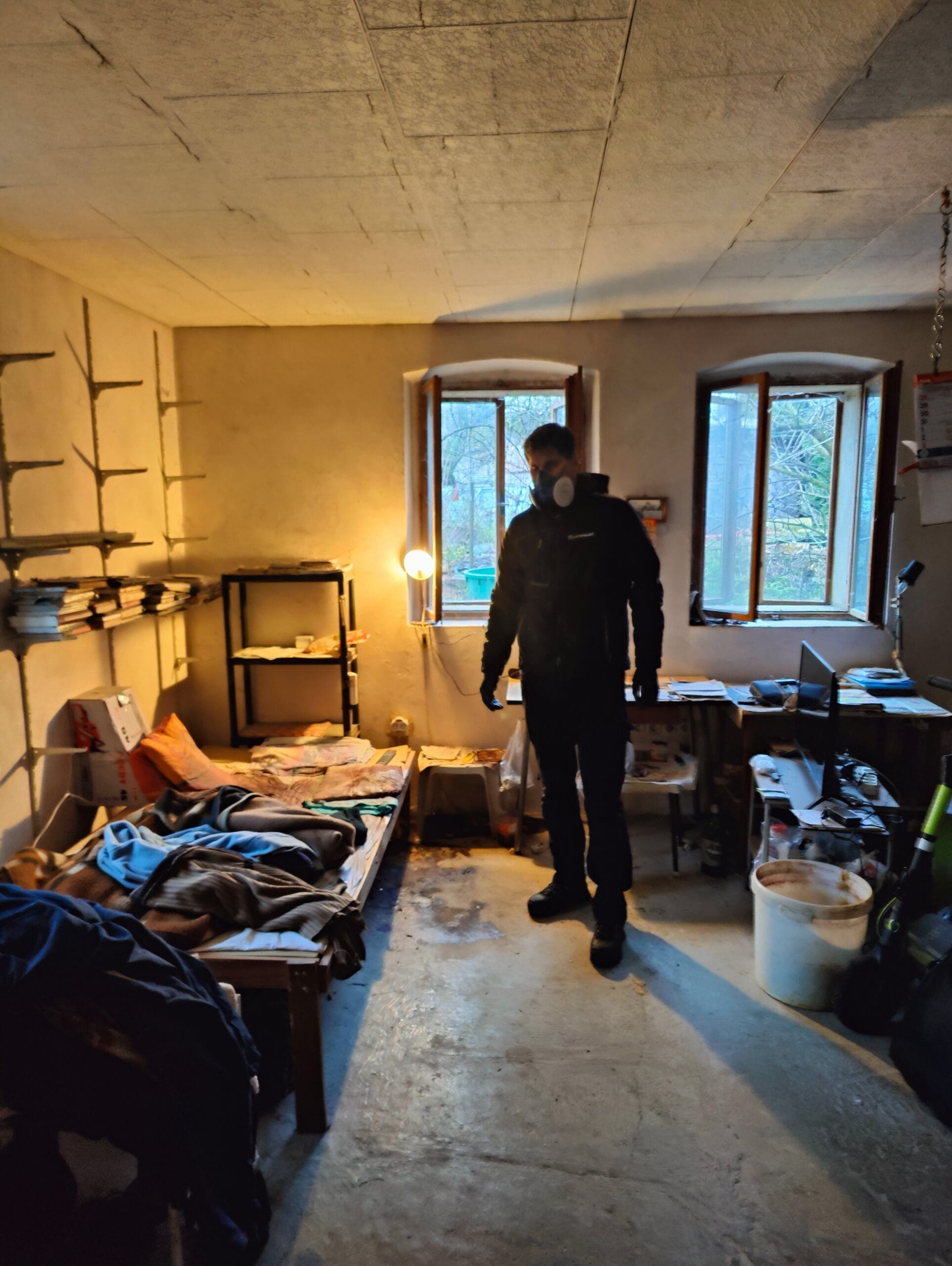 Muž v ochranném oděvu při vyklízení sklepa s rozházenými věcmi.