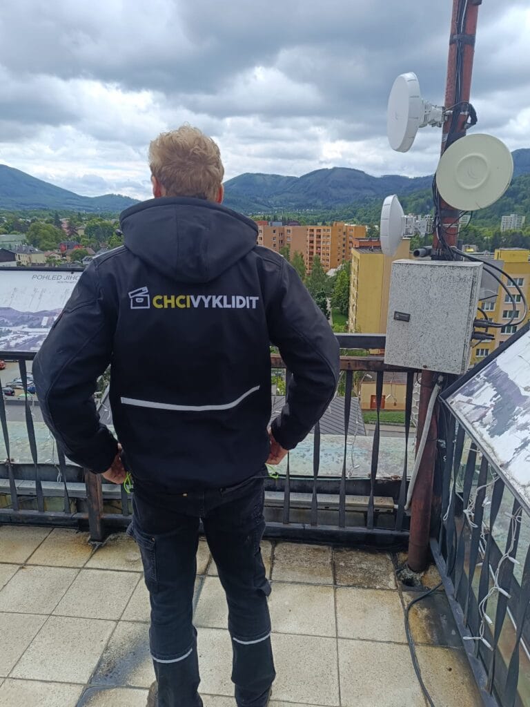 Muž v kabátě s logem ChciVyklidit.cz na střechě s výhledem na město a hory.